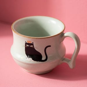 Puede incluir: Una taza de cerámica gris claro con cuerpo redondeado y asa curva. La taza presenta una ilustración gráfica de un gato negro con patas blancas y un pequeño detalle naranja en la cabeza, situada en la parte frontal del recipiente.