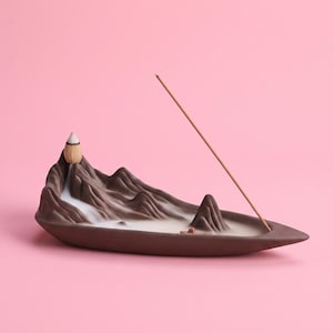 Peut inclure: Un brûleur d'encens en céramique brune en forme de chaîne de montagnes avec une base en forme de bateau. Un cône d'encens allumé repose sur le sommet, libérant une fumée blanche qui descend le long des pentes, tandis qu'un bâtonnet est incliné.
