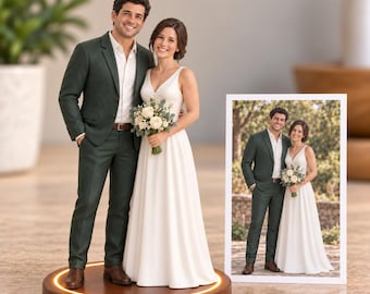 May include: A custom wedding cake topper figurine set featuring a person in a dark green suit and a person in a white wedding dress holding a bouquet, standing on a circular wooden base with a light ring, next to a framed photograph of the couple.