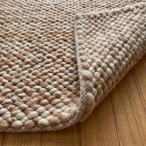 May include: A thick, hand-woven area rug featuring a chunky, textured knit pattern in alternating shades of cream, beige, and light brown. The rug is folded at the corner, revealing a dense, braided construction and a finished, reinforced edge.