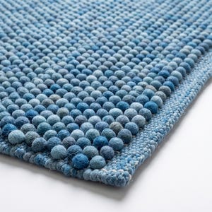 May include: A rectangular rug constructed from numerous small, spherical felted wool balls in varying shades of blue, ranging from light sky blue to deep navy. The surface features a dense, textured pattern with a flat, woven border edge.