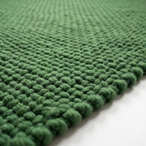 May include: A thick, forest green rug featuring a dense, hand-knitted texture with prominent, uniform loops. The material displays a consistent deep green hue throughout, showcasing a chunky, tactile surface pattern that extends across the entire visible area.