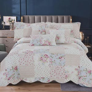 May include: A patchwork quilt and matching pillow shams featuring a floral pattern with pink roses and small blue flowers on a white background. The bedding set has a scalloped edge and is displayed on a bed with a grey headboard.
