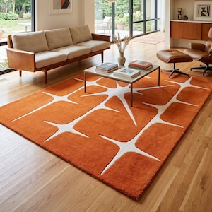 May include: A rectangular area rug featuring a vibrant orange base color with a repeating white starburst or geometric cross pattern. The rug has a plush, textured surface and is positioned on a light-colored wooden floor in a living room setting.