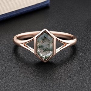 May include: A rose gold ring featuring a hexagonal moss agate gemstone set in a split shank band. The stone displays translucent green and grey inclusions within its structure. The metal band has a polished finish and a geometric design.