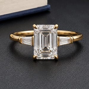 May include: A gold ring featuring a large emerald-cut center stone flanked by two smaller tapered baguette-cut side stones. The band is polished yellow gold, and the setting holds the three clear, faceted stones securely with metal prongs.