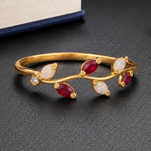 Puede incluir: Un anillo de oro con una banda delicada en forma de enredadera, adornada con rubíes rojos y ópalos blancos alternados en talla marquesa, acentuado por una pequeña piedra preciosa redonda y transparente en un extremo, con acabado pulido.