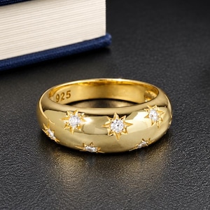 May include: A gold-toned band ring featuring multiple star-shaped engravings, each centred with a small clear stone. The interior of the band is clearly stamped with the number 925, indicating sterling silver composition with a gold-coloured finish.