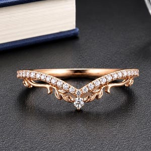 May include: A rose gold V-shaped ring featuring a series of small, clear, round-cut stones set along the band. The metal band is crafted with intricate, leaf-like filigree detailing that extends from the centre point outward along the sides.