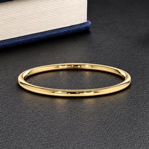 May include: A thin, polished gold band ring with a smooth, rounded surface. The ring features a simple, circular design with a reflective metallic finish that catches the light, showcasing a clean and uniform appearance throughout the entire piece.