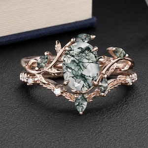 Peut inclure: Une bague en or rose ornée d'une pierre centrale en agate mousse de taille ovale avec des inclusions vert foncé. L'anneau présente un motif de branches, agrémenté de petites pierres d'agate mousse taille marquise et de minuscules pierres claires.