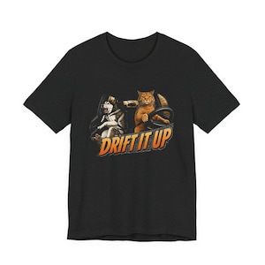 May include: A black short-sleeve t-shirt featuring a graphic print of a husky and an orange tabby cat sitting in the front seats of a car. The cat is holding the steering wheel. Below the animals, the text reads DRIFT IT UP in orange letters.