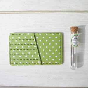 May include: A green polka dot card holder with a stitched design and a small glass vial with a cork stopper. The card holder is rectangular and has white polka dots. The vial contains embroidery needles.