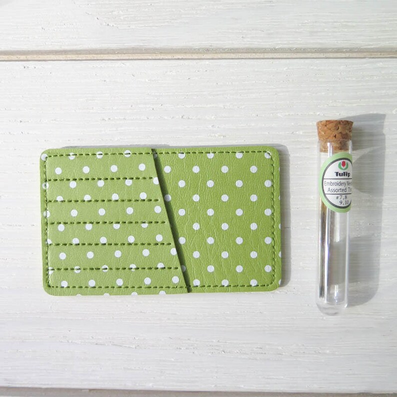 May include: A green polka dot card holder with a stitched design and a small glass vial with a cork stopper. The card holder is rectangular and has white polka dots. The vial contains embroidery needles.