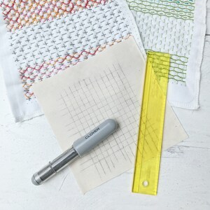 May include: A craft image featuring a gray Clover marking pen, a yellow ruler, and fabric samples with stitched patterns. The fabric samples display colorful embroidery and grid lines, suggesting a sewing or quilting project.