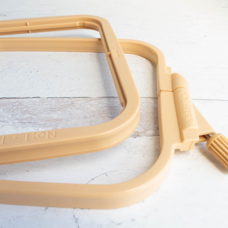 May include: Two beige embroidery hoops, one partially assembled, are displayed against a light wood-grain background. The hoops are made of plastic and feature a screw for tension adjustment. The text "No. 1 - 1" is visible on one hoop, and "65 mm" on another.