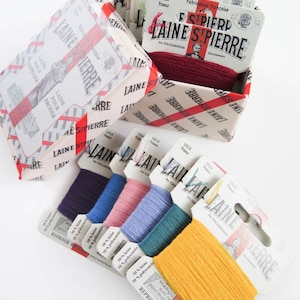 May include: A collection of colorful embroidery floss in various shades, including blue, pink, and yellow. The floss is packaged with labels that read "LAINE ST PIERRE" and "LILLE France". The image also shows a box with the same branding.