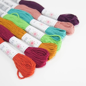 May include: A collection of colorful embroidery floss skeins in shades of orange, red, green, teal, and purple. Each skein is labeled with a white paper band featuring a floral design. The threads are arranged on a white surface.