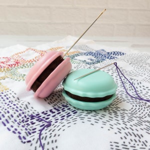 May include: Two macaron-shaped needle minders in pink and teal, each with a dark brown filling. A gold needle is inserted into each minder. A purple thread extends from the teal minder. The items rest on a white cloth with colorful embroidery.