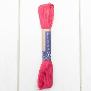 Puede incluir: Madeja de hilo de bordar rosa vibrante con una etiqueta de papel. La etiqueta presenta texto japon&eacute;s y un dise&ntilde;o azul y dorado. La madeja mide aproximadamente 40 metros de largo y se muestra sobre un fondo de madera blanca.