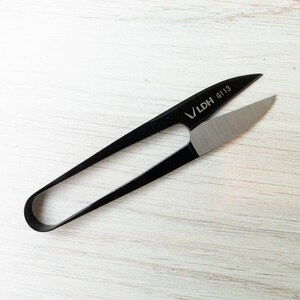 May include: Black LDH G113 thread snips with silver blades, lying on a white wooden surface. The snips have a curved handle and pointed blades, designed for precision cutting of threads and small materials.