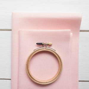 May include: A wooden embroidery hoop with a black and gold metal clasp rests on a stack of light pink cross-stitch fabric. The fabric is neatly folded, showing its woven texture. The hoop is round and made of light-colored wood.