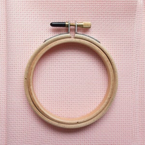 May include: A wooden embroidery hoop with a brass screw and black adjustment knob, set against a light pink cross-stitch fabric. The hoop is round, with a light wood grain, and is designed to hold fabric taut for needlework projects.