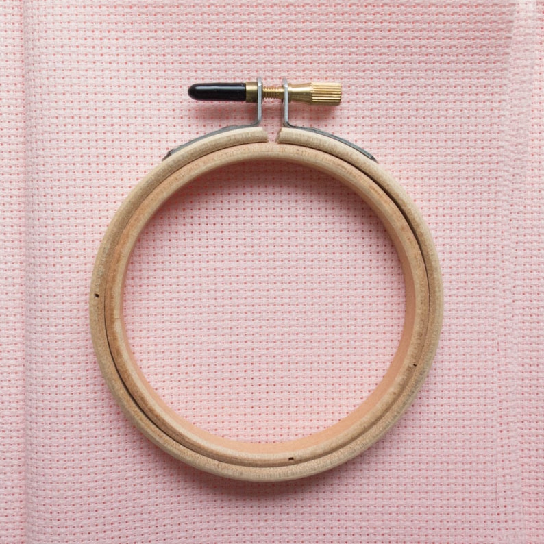 May include: A wooden embroidery hoop with a brass screw and black adjustment knob, set against a light pink cross-stitch fabric. The hoop is round, with a light wood grain, and is designed to hold fabric taut for needlework projects.