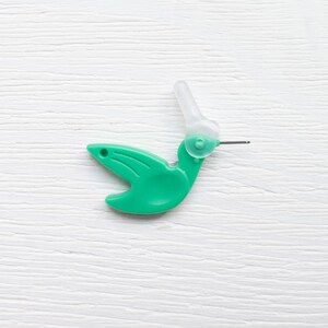 May include: A green plastic hummingbird-shaped needle threader with a clear plastic section and a thin metal wire. The threader is designed to assist in threading needles. The design features a bird with a curved beak and wings.