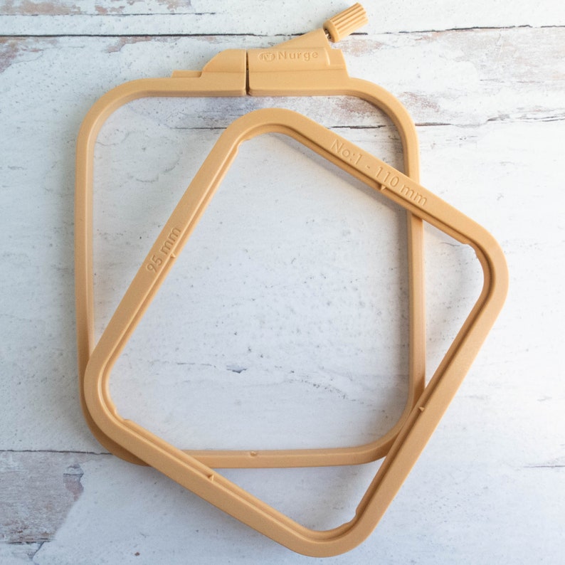 May include: Two beige square embroidery hoops, one open and one closed. The closed hoop has a screw adjustment and the brand name "Nurge". The open hoop is labeled "95 mm" and the other "No. 1 - 110 mm".