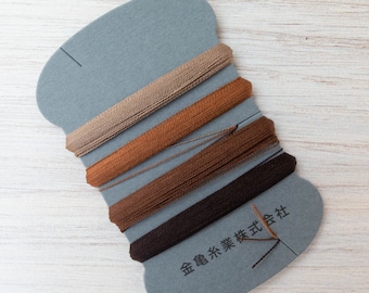 May include: A spool of thread in shades of brown, from light tan to dark chocolate. The thread is neatly wound on a gray, stylized card with Japanese text.