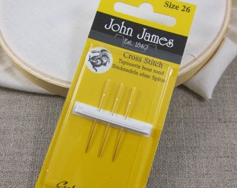 May include: A package of gold-plated cross-stitch needles from John James, size 26. The yellow and black packaging displays the brand name, needle count, and product details. The needles are designed for tapestry and cross-stitch projects.