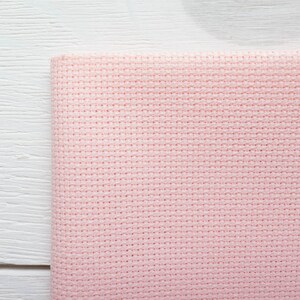May include: A folded piece of light pink cross-stitch fabric. The fabric has a grid-like pattern of evenly spaced holes, ideal for needlework. The fabric is set against a white wooden surface.