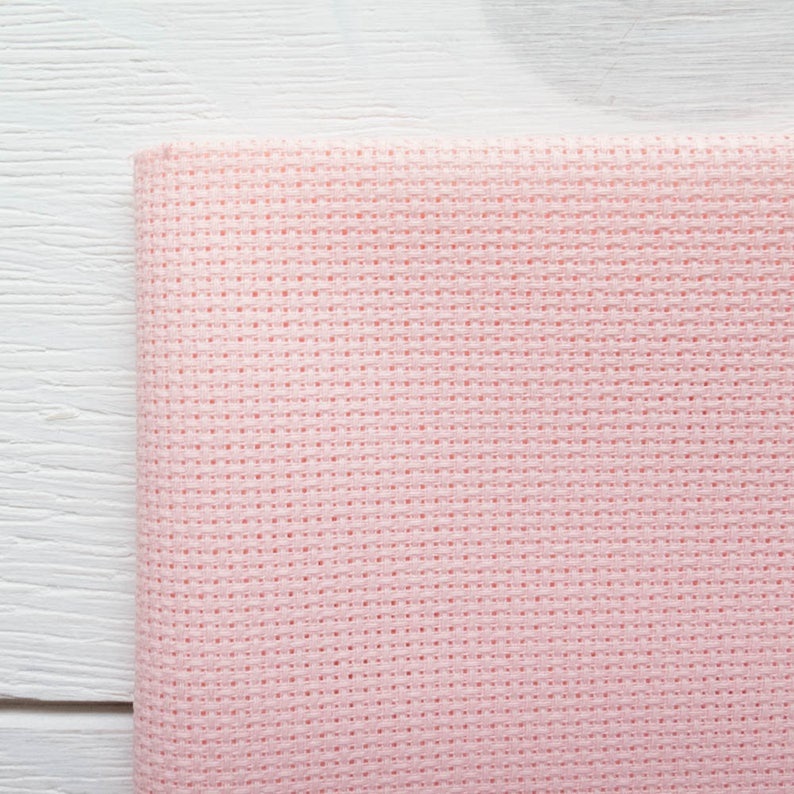 May include: A folded piece of light pink cross-stitch fabric. The fabric has a grid-like pattern of evenly spaced holes, ideal for needlework. The fabric is set against a white wooden surface.