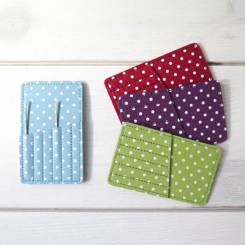 May include: A collection of colorful, polka-dotted needle cases and holders. The cases are in shades of blue, red, purple, and green, with white polka dots. The blue case holds two needles. The cases are arranged on a white wooden surface.