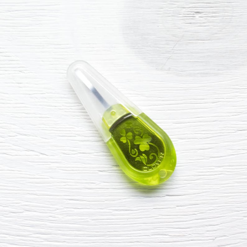 May include: A lime green seam ripper with a clear plastic safety cap. The ripper has a clover design etched into the green plastic. The tool is designed for removing stitches from fabric.