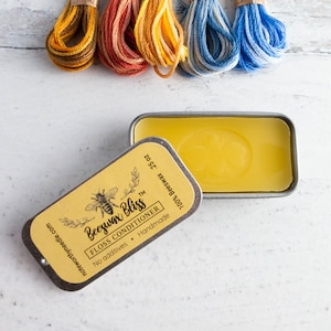 May include: A tin of beeswax floss conditioner with the text "Beeswax Bliss" and a bee illustration, next to a tin containing a yellow wax block. Colorful embroidery floss in shades of yellow, orange, and blue are in the background.
