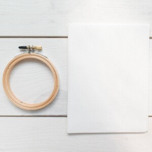 May include: A wooden embroidery hoop with a metal clasp and a white piece of Aida cloth. The hoop is round and made of light-coloured wood. The cloth is rectangular and has a visible grid pattern.