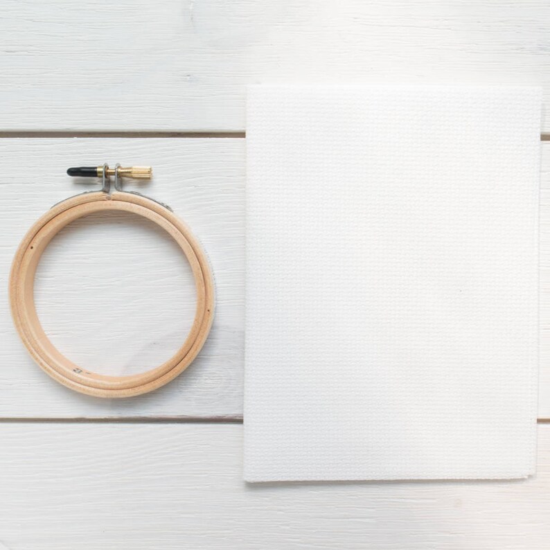 May include: A wooden embroidery hoop with a metal clasp and a white piece of Aida cloth. The hoop is round and made of light-coloured wood. The cloth is rectangular and has a visible grid pattern.