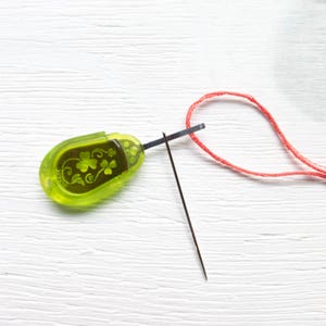 May include: A green needle threader with a clover design and a black needle. A red thread is looped through the needle. The threader is made of plastic and has the word "JAPAN" printed on it.