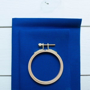 May include: A wooden embroidery hoop with a brass screw is clamped onto a piece of blue fabric. The fabric is a solid, deep blue colour and has a textured appearance. The hoop is round and made of light-coloured wood.