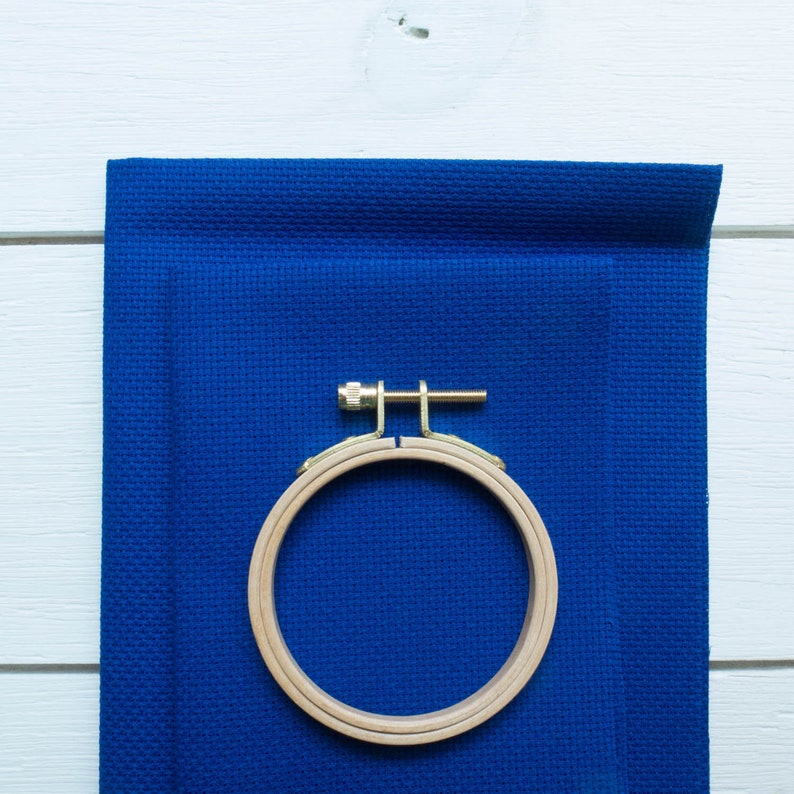May include: A wooden embroidery hoop with a brass screw is clamped onto a piece of blue fabric. The fabric is a solid, deep blue colour and has a textured appearance. The hoop is round and made of light-coloured wood.