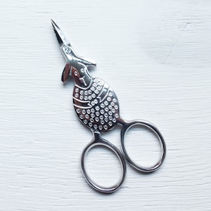 May include: Silver embroidery scissors with a bird motif. The scissors have a detailed bird body with a textured pattern and a long, pointed beak. The handles have two finger holes. The scissors are set against a white wooden background.