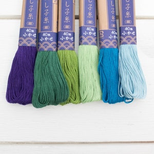 May include: Six skeins of embroidery floss in shades of purple, green, and blue. Each skein is labeled with Japanese text and contains 40 meters of thread. The floss is arranged on a white wooden surface.