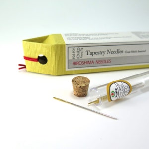 May include: A yellow box of Hiroshima Tapestry Needles with a red string closure. A cork stopper and a clear tube of needles are next to the box. The text on the box reads "Tapestry Needles Cross Stitch Assorted".