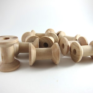 May include: A collection of unfinished wooden spools, perfect for crafts. The spools are cylindrical with a central hole and flared ends. The natural wood grain is visible, and the spools are arranged on a white surface.