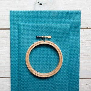 May include: A wooden embroidery hoop with a brass screw is placed on top of two pieces of teal cross-stitch fabric. The fabric has a textured appearance. The hoop is approximately 6 inches in diameter. The background is a white wooden surface.