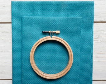 May include: A wooden embroidery hoop with a brass screw is placed on top of two pieces of teal cross-stitch fabric. The fabric has a textured appearance. The hoop is approximately 6 inches in diameter. The background is a white wooden surface.