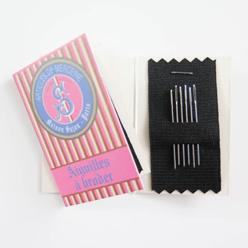 May include: A sewing needle set with a pink and white striped box featuring the text "ARTICLES DE MERCERIE Maison Sajou - Paris" and "Aiguilles &agrave; broder". The set includes multiple silver needles on a black fabric.