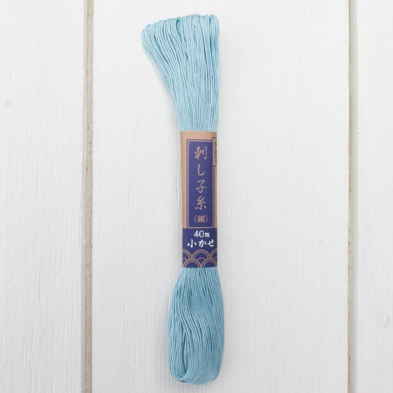 May include: A skein of light blue embroidery floss with a brown label. The label has Japanese text and reads "40m". The floss is neatly bundled and ready for use. The background is a white wooden surface.